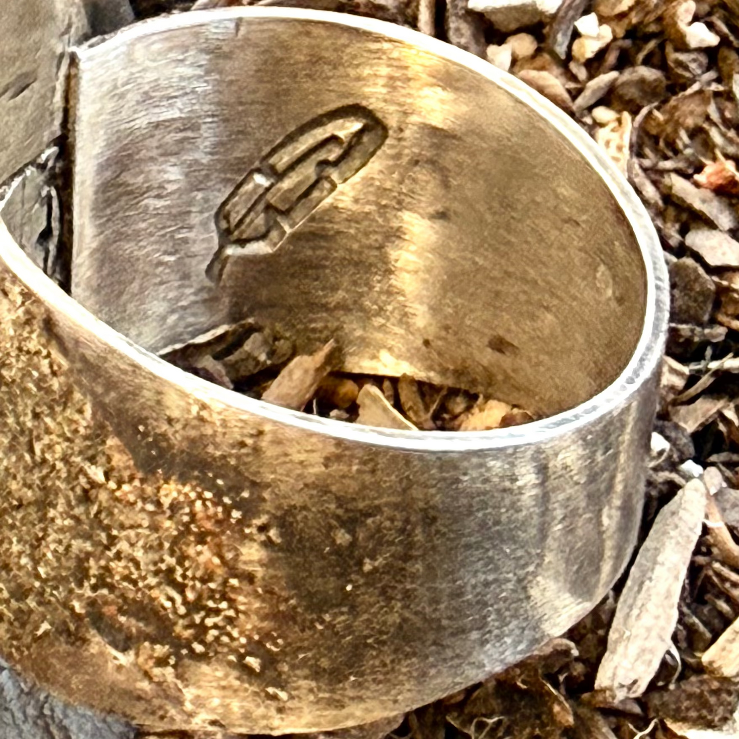 A wide, chunky silver ring with a textured exterior and a polished inner band, shown lying on a bed of wood chips and small stones. Stamped feather on inside of band.