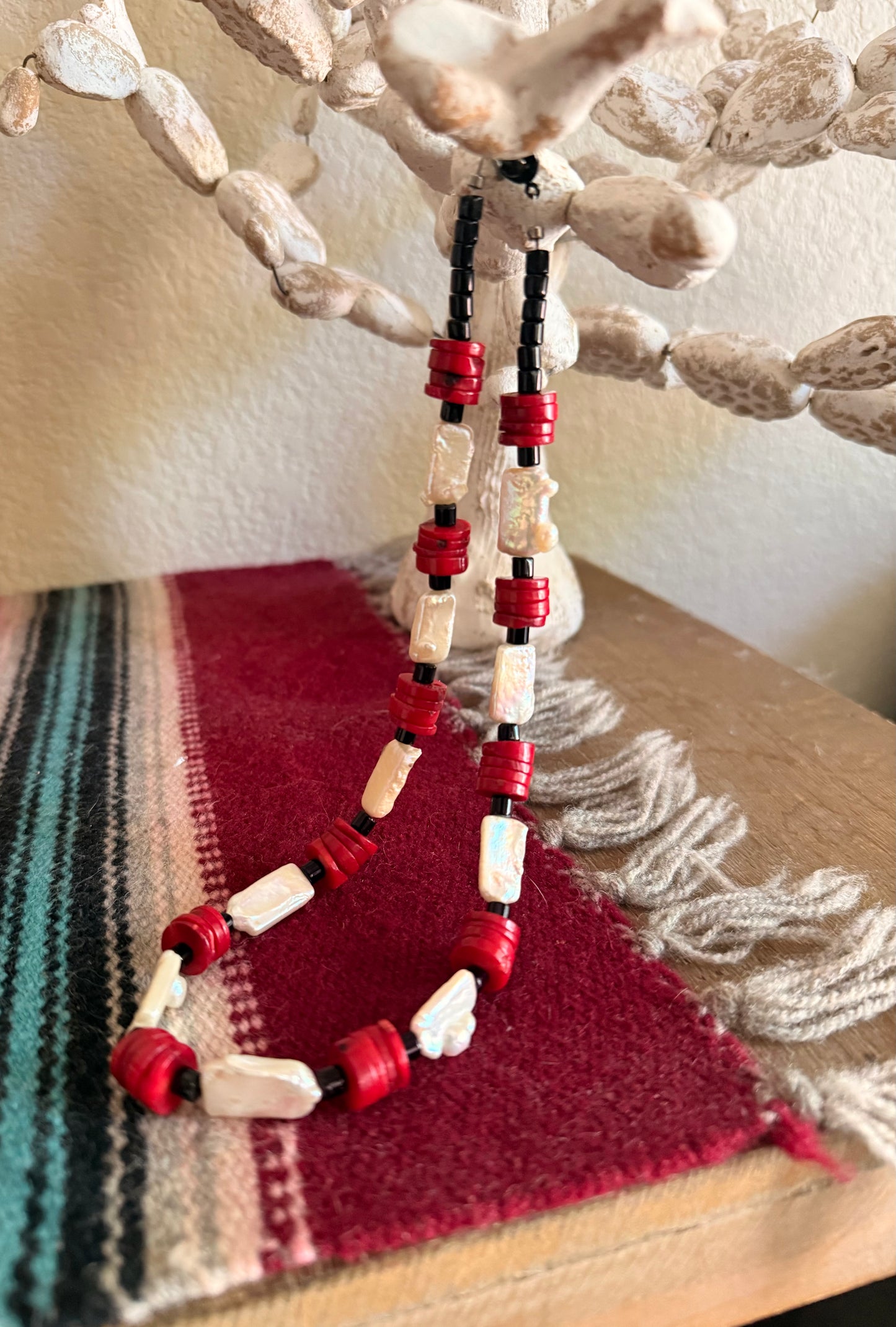 A handmade beaded necklace with red cylindrical coral beads, white shell beads, and black spacer onxy beads, displayed on a wooden stand with a red and green woven textile underneath.