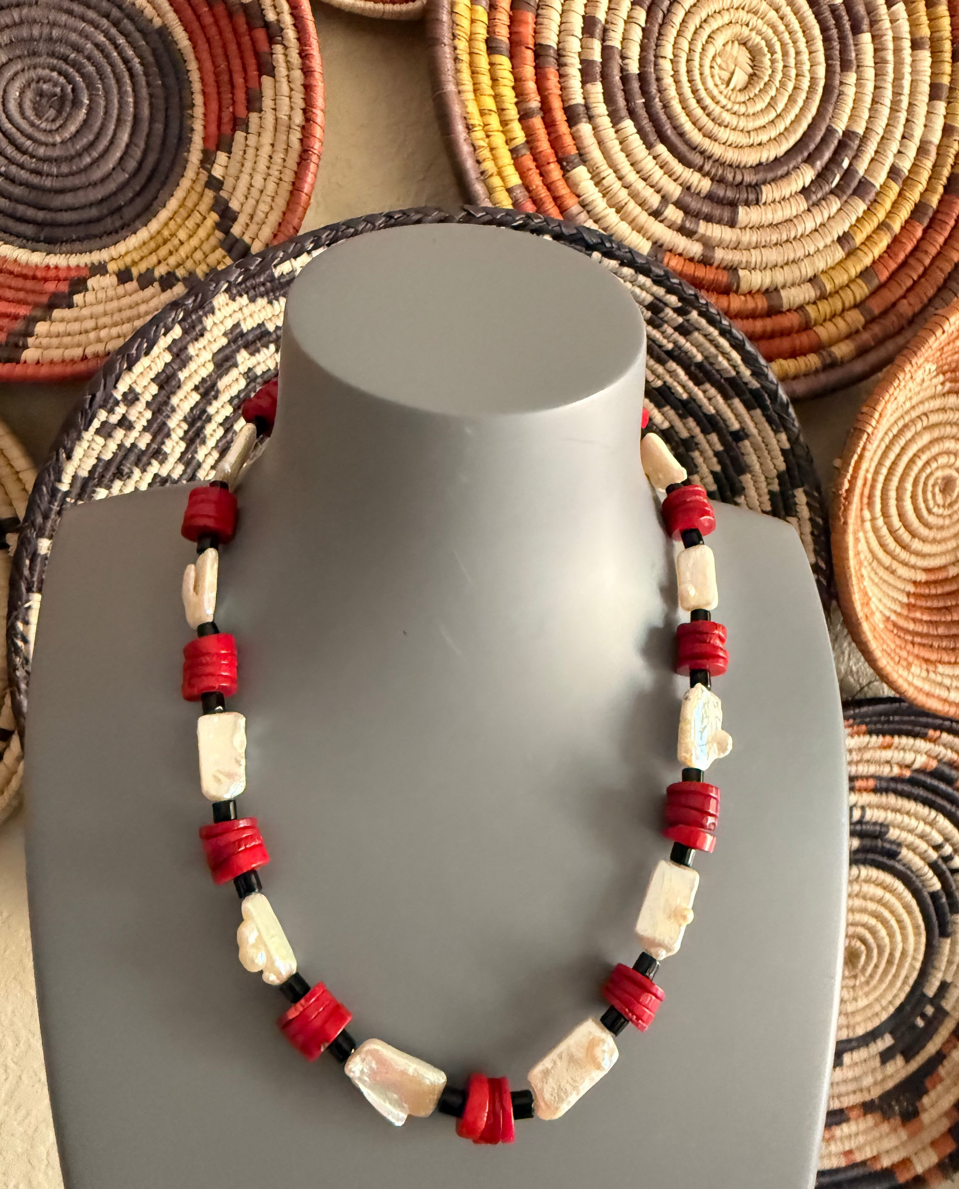 A beaded necklace displayed on a gray mannequin bust, featuring red coral cylindrical beads, white rectangular baroque fresh water pearl beads, separated by small black onyx spacer beads. The background shows woven circular baskets in earthy tones.